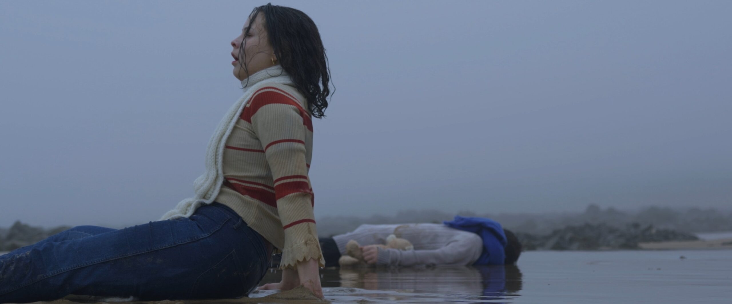 Isalys tente de se relever sur la plage détrempée tandis que son frère Élio gît à ses côtés, illustrant le combat pour la vie dans Entre deux eaux.