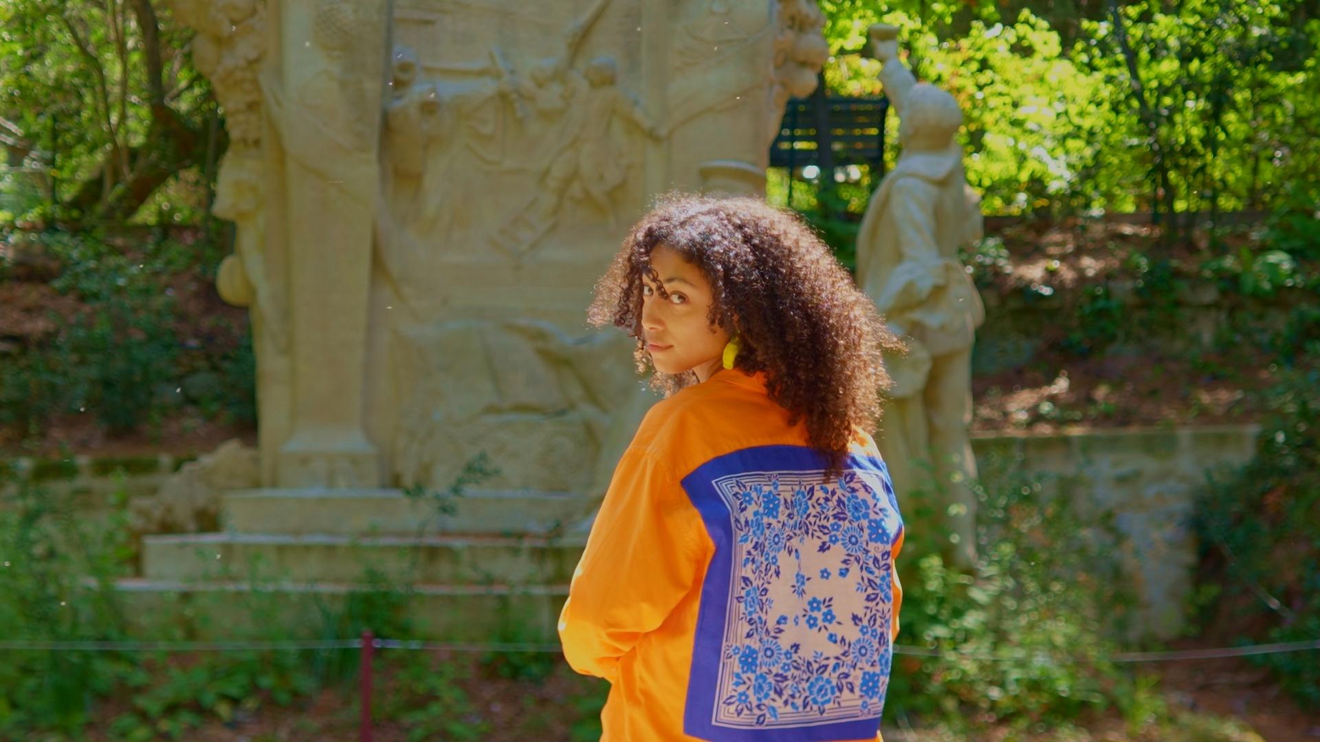 Portrait d'une femme portant une veste orange à motifs bleus, prise devant une sculpture en plein air pour le projet Maison Janie, une marque de mode éthique et méditerranéenne mise en lumière par O3il Production.