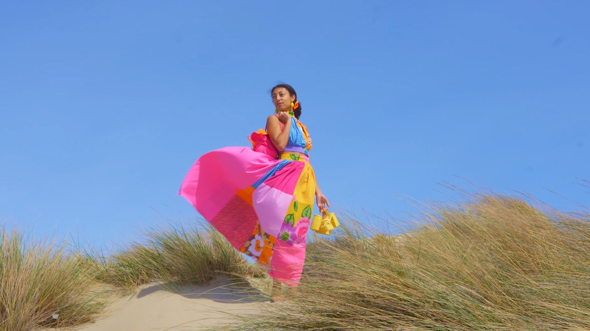 Femme vêtue d’une robe colorée Maison Janie posant sur une dune sous un ciel bleu, image captée par O3il Production.